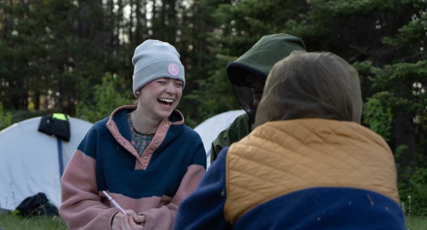 Three students sit in a wooded area. The one whose face we can see is laughing. There are tents in the background. 